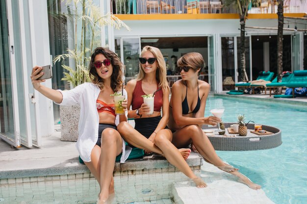 Speelse brunette vrouw in zonnebril selfie maken met vrienden in het resort. Gebruinde blanke vrouwen die een foto van zichzelf in het zwembad nemen.