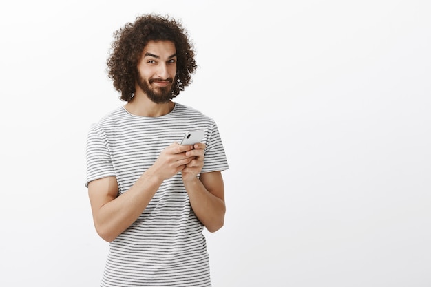 Speelse, doordachte knappe man met krullend haar, die smartphone vasthoudt, grijnzend en kijkt met een nieuwsgierige, geïntrigeerde uitdrukking