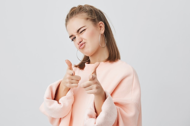 Speelse jonge blanke vrouw met recht blond haar in roze sweatshirt met lange mouwen, staand, spottend, wijzend met haar wijsvingers naar jou