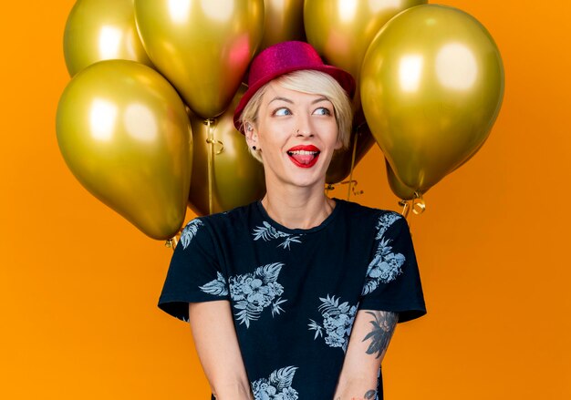 Speelse jonge blonde partijvrouw die partijhoed draagt die zich voor ballons bevindt die kant bekijkt die tong toont die op oranje muur wordt geïsoleerd