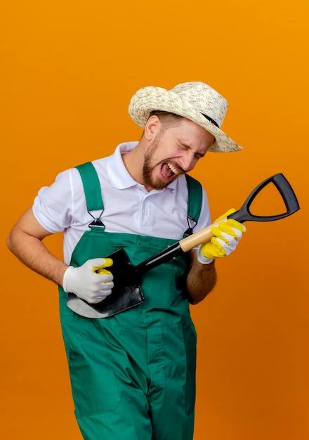 Gratis foto speelse jonge knappe slavische tuinman in uniform dragen hoed en tuinhandschoenen houden schop doen alsof gitaar spelen met gesloten ogen geïsoleerd op oranje muur