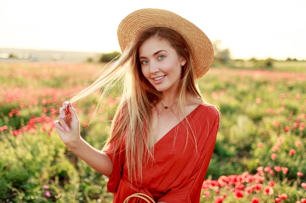 Speelse, onbezorgde blonde vrouw die graag poseren tijdens een wandeling buiten in een gelukkige stemming. Met strooien hoed, oranje playsuit. Papaver veld.