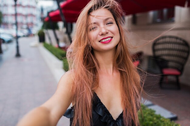 Speelse schattige rode hoort vrouw met glimlachen zelfportret maken en genieten van zomervakantie in Europa. Positief buitenbeeld. Zwarte jurk, rode lippen.
