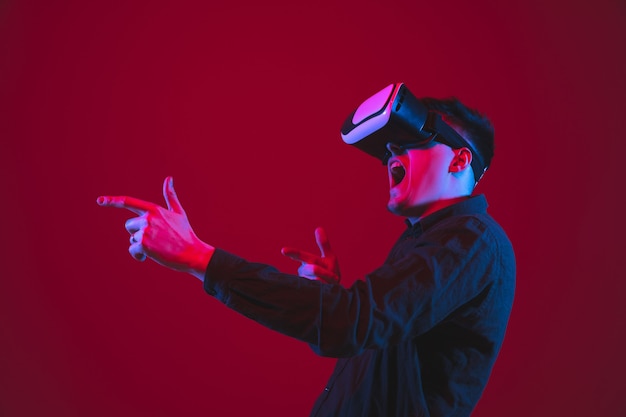 Spelen droeg een VR-headset. Portret van een blanke jongeman geïsoleerd op rode muur in neonlicht. Mooi modelletje. Concept van menselijke emoties, gezichtsuitdrukking, jeugd, apparaten.