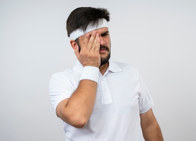 Spijt jonge sportieve man kijken kant dragen hoofdband en polsband bedekt oog met hand geïsoleerd op een witte muur