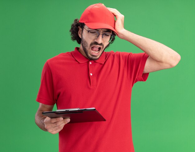 Spijt van jonge bezorger in rood uniform en pet met een bril met klembord en potlood die hand op het hoofd zetten en kijken naar klembord geïsoleerd op groene muur