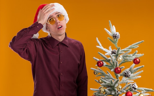 Gratis foto spijt van jonge blonde man met kerstmuts en bril permanent in de buurt van versierde kerstboom opzoeken houden hand op hoofd geïsoleerd op een oranje achtergrond