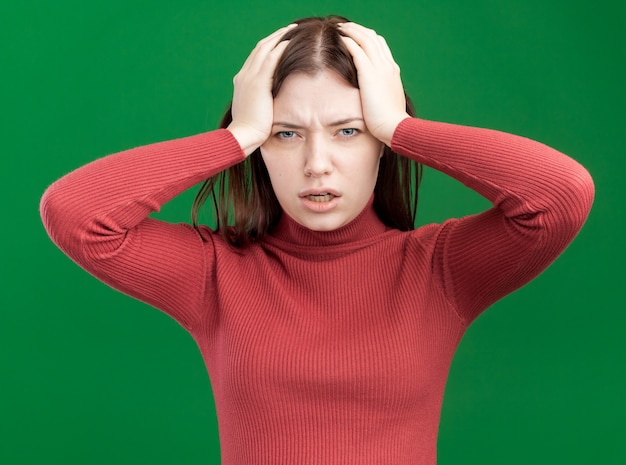 Spijt van jonge mooie vrouw die naar de voorkant kijkt en de handen op het hoofd houdt geïsoleerd op de groene muur