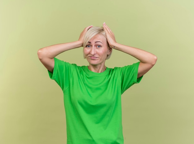 Gratis foto spijt van middelbare leeftijd blonde slavische vrouw die voorzijde bekijkt die handen op hoofd houdt die op olijfgroene muur wordt geïsoleerd