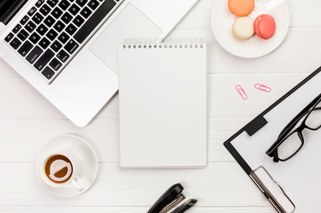 Spiraalvormige blocnote met laptop, makarons, koffiekop met klembord en oogglazen op wit bureau