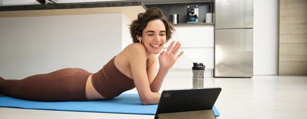 Gratis foto sport en levensstijl jonge fitnessvrouw gyminstructeur zwaait met de hand op de tablet en sluit zich aan bij de online videoklasse