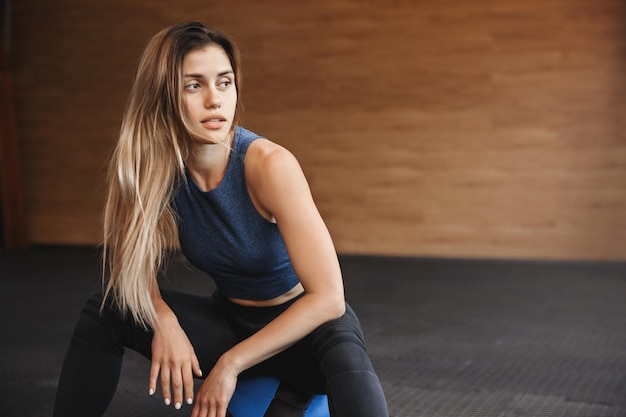 Sport fitness en uithoudingsvermogen concept prachtige jonge fitness instructeur sportvrouw in activewear met lang blond haar zit medicijnbal in de buurt van houten sportschool muur zet rust opzij voor de training