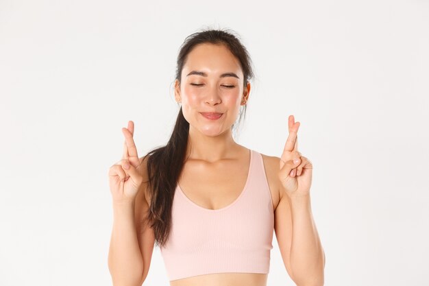 Sport, welzijn en actief levensstijlconcept. Close-up van glimlachend optimistisch Aziatisch meisje, hoop afvallen, kruis vingers voor geluk en ogen sluiten tijdens het doen van wens, witte muur