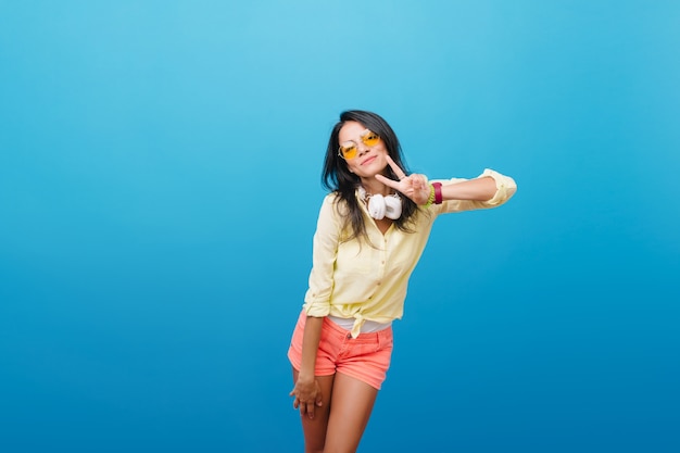 Sportief gelooid meisje in denim shorts poseren met plezier. Indoor portret van prachtige Europese vrouw in straat outfit draagt grote koptelefoon.