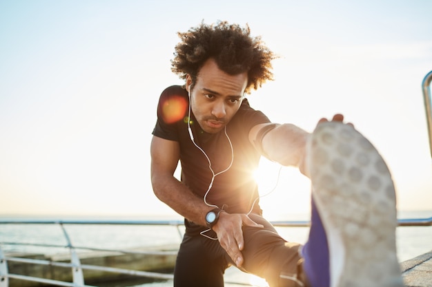 Sportieve Afro-Amerikaanse jongen in zwarte sportkleding en blauwe sneakers die zijn spieren strekt