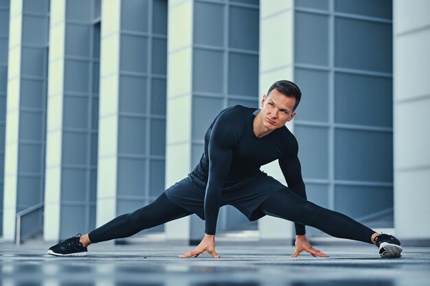 Sportieve atletische man warmt zich op en strekt zich buiten uit over de achtergrond van een modern gebouw.