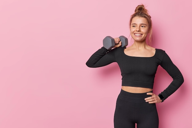 Sportieve fit jonge vrouw heeft goed ontwikkelde spieren door krachttraining verhoogt halter heeft training in sportschool houdt hand op taille poses tegen roze achtergrond blanco kopieerruimte voor uw promotie