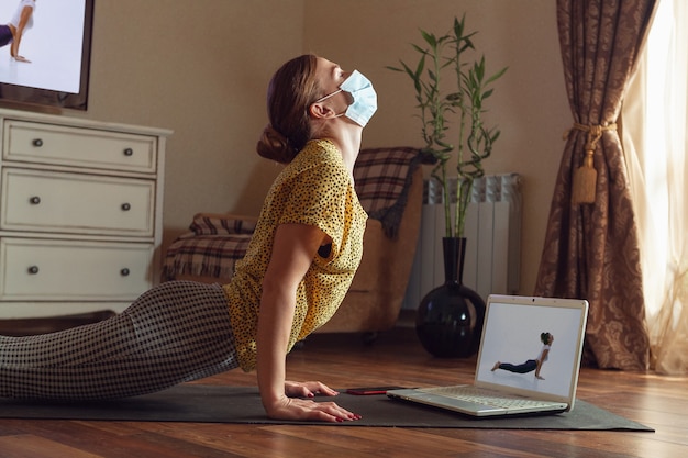 Sportieve jonge vrouw die online yogalessen volgt en thuis oefent terwijl ze in quarantaine is. Concept van een gezonde levensstijl, wellness, veilig zijn tijdens een pandemie van het coronavirus, op zoek naar een nieuwe hobby.