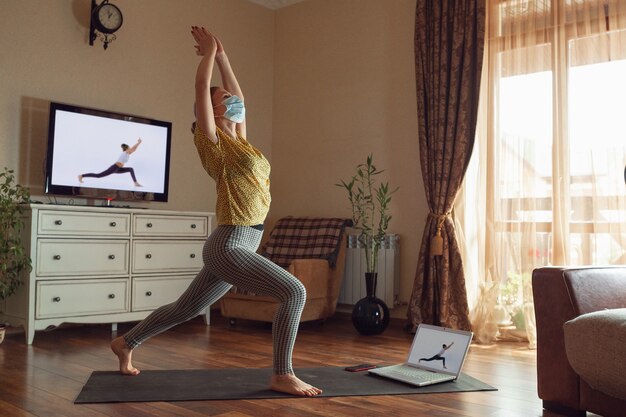 Sportieve jonge vrouw die online yogalessen volgt en thuis oefent terwijl ze in quarantaine is. Concept van een gezonde levensstijl, wellness, veilig zijn tijdens een pandemie van het coronavirus, op zoek naar een nieuwe hobby.