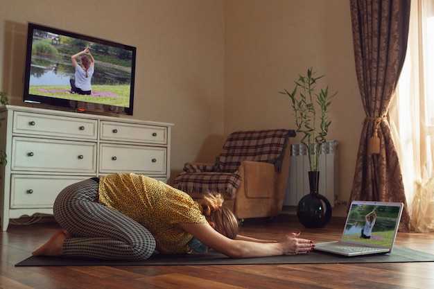Sportieve jonge vrouw die online yogalessen volgt en thuis oefent terwijl ze in quarantaine is. Concept van een gezonde levensstijl, wellness, veilig zijn tijdens een pandemie van het coronavirus, op zoek naar een nieuwe hobby.