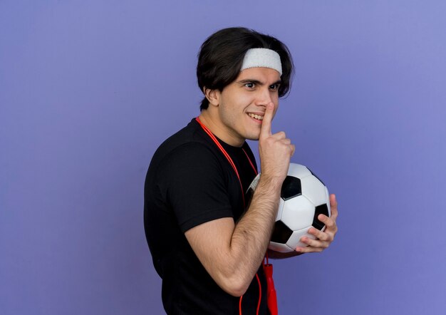 Sportieve jongeman met sportkleding en hoofdband met springtouw rond nek houden voetbal stilte gebaar met vinger op lippen maken