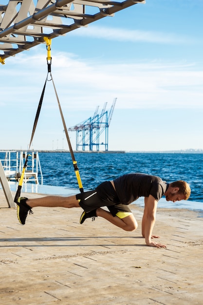 Gratis foto sportieve jongeman training met trx in de buurt van de zee in de ochtend.