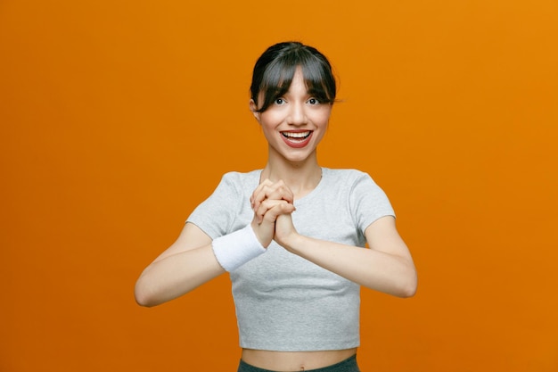 Sportieve mooie vrouw in sportkleding die naar de camera kijkt die haar handen uitrekt voordat ze gaat trainen, glimlachend zelfverzekerd over een oranje achtergrond