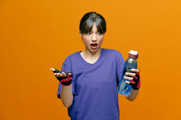 Sportieve, mooie vrouw in sportkleding in handschoenen die een fles water vasthoudt en ernaar kijkt dat ze verward en bezorgd over een oranje achtergrond staat