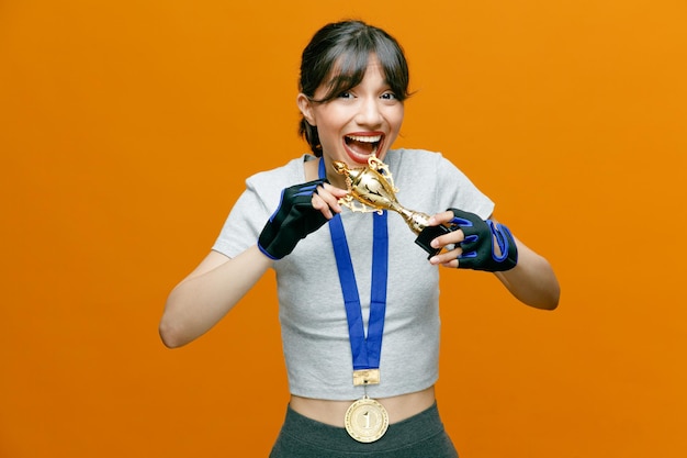 Sportieve mooie vrouw in sportkleding in handschoenen met winnende medaille om haar nek met trofee kijkend naar camera blij en opgewonden blij met haar succes over oranje achtergrond