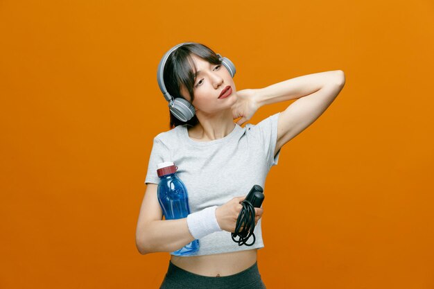 Sportieve mooie vrouw in sportkleding met koptelefoon op haar hoofd met springtouw en fles water die er moe uitziet, houdt de hand op haar nek over oranje achtergrond