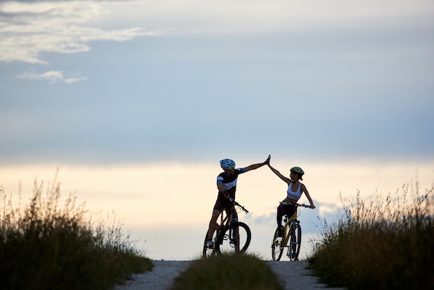 Sportieve vrouw en man berijdende fietsen, die pret hebben buiten. silhouetten van sportmannen die vijf verhogen en op weg in zonsondergangtijd stellen. niet-stedelijke scene.