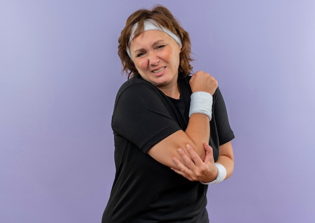 Gratis foto sportieve vrouw van middelbare leeftijd in zwart t-shirt met hoofdband die onwel kijkt wat betreft haar elleboog die pijn voelt die zich over blauwe muur bevindt