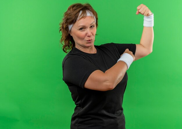 Sportieve vrouw van middelbare leeftijd in zwart t-shirt met hoofdband die vuist opheft die biceps toont die zelfverzekerd en gelukkig status over groene muur kijken