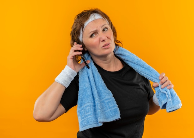 Sportieve vrouw van middelbare leeftijd in zwart t-shirt met hoofdband en handdoek op schouder op zoek moe praten op mobiele telefoon staande over oranje muur