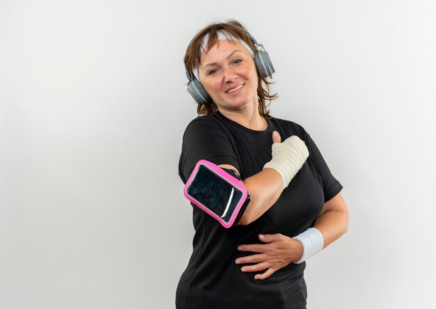 Sportieve vrouw van middelbare leeftijd in zwart t-shirt met hoofdband en smartphone armband hand op haar borst houden dankbaar gevoel staande over witte muur