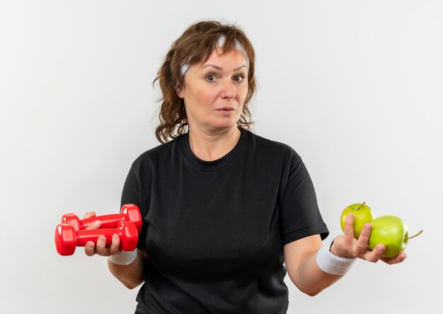 Sportieve vrouw van middelbare leeftijd in zwart t-shirt met hoofdband met twee groene appels en halters op zoek verward met twijfels staande over witte muur