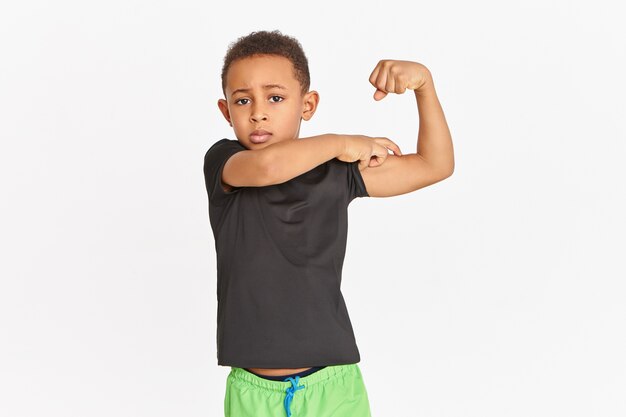 Sportieve zelfverzekerde Afrikaanse jongen in sportkleding die biceps aanspant, kracht en fysiek uithoudingsvermogen aantoont. Leuk atletisch kind met een donkere huid dat trots is op zichzelf en pronkt met zijn gespannen armspieren