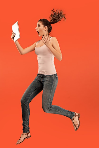 Sprong van jonge vrouw over blauwe studioachtergrond die tabletgadget gebruikt tijdens het springen.