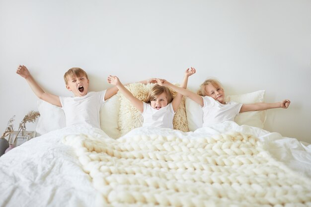 Sta op en schijn. Geïsoleerde horizontale opname van drie broers en zussen, zusje en haar twee oudere broers die identieke witte t-shirts dragen, zittend op bed, armen strekken en geeuwen in de ochtend