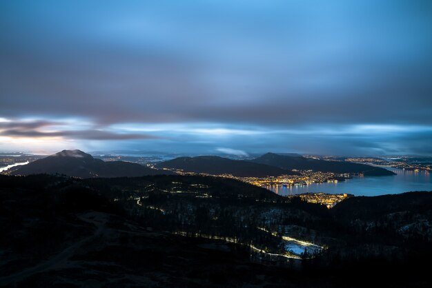 Stad omgeven door bergen en een zee bedekt met lichten onder een bewolkte hemel in de avond