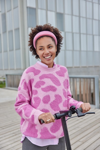 Stadsleven concept positief millennial meisje rijdt elektrische kick scooter draagt hoofdband jumper in stadsgezicht beweegt door de stad kijkt graag in de verte krullendharige vrouw gebruikt ecologisch vervoer