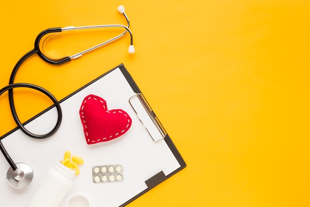 Stethoscoop; gestikt hart; medicijnen die uit flessen vallen; blisterverpakking geneeskunde met klembord over gele tafel