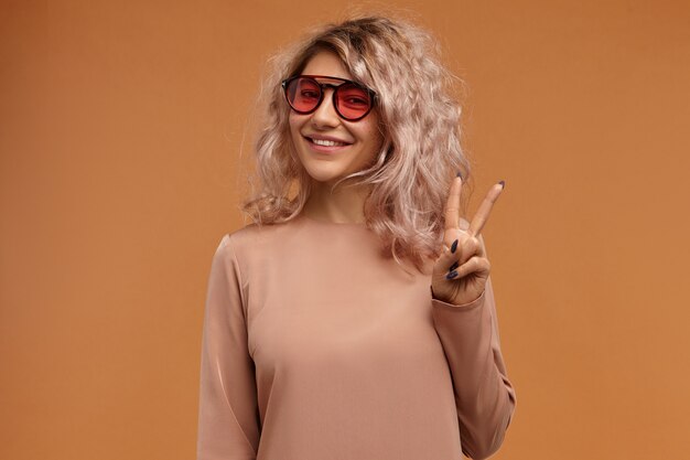 Stijl, modetrends en jeugdconcept. Foto van modieus hipster meisje trendy zonnebril met roze lenzen met vrolijke brede glimlach, vredesteken maken
