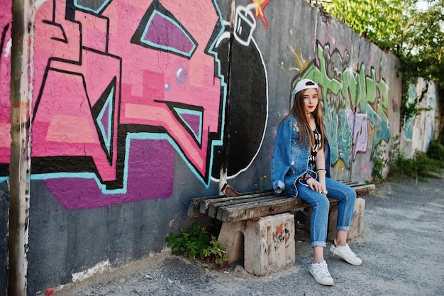 Stijlvol, casual hipster-meisje in pet en spijkerbroek draagt luistermuziek van een koptelefoon van een mobiele telefoon tegen een grote graffitimuur met bom