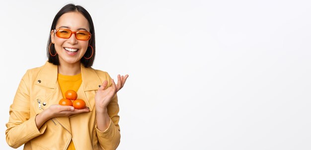 Stijlvol gelukkig Aziatisch meisje in zonnebril met mandarijnen en glimlachend poseren tegen een witte achtergrond