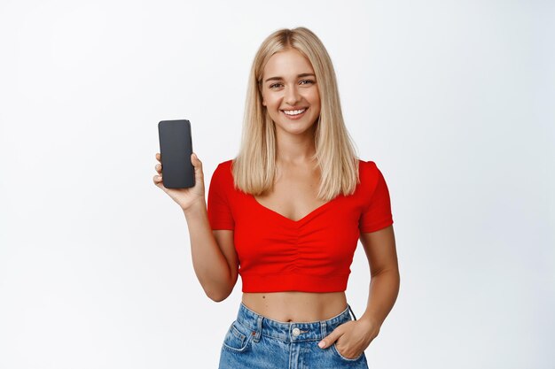 Stijlvol glimlachend blond meisje dat het smartphonescherm toont en een mobiele app aanbeveelt die op een witte achtergrond staat