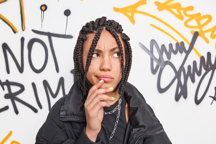 Gratis Foto stijlvol hipster meisje heeft dreadlocks diep in gedachten houdt hand in de buurt van mond geconcentreerd boven poses in stedelijke omgeving tegen graffitimuur draagt zwarte jas