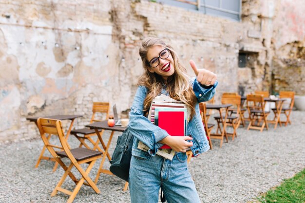 Stijlvol hipster meisje in het retro jeans pak poseren voor de oude bakstenen muur. Trendy jonge vrouw met zak die zich naast oud gebouw bevindt.