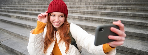 Gratis foto stijlvol jong meisje met rode hoed maakt foto's op de smartphonecamera en maakt selfie terwijl ze op de trap zit