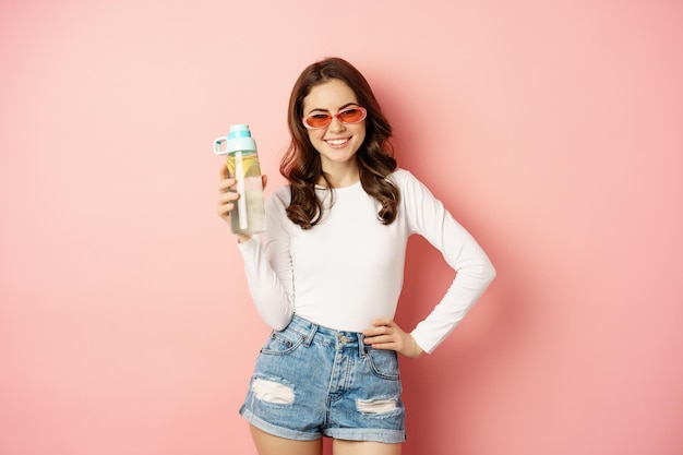 Stijlvol meisje in lente-outfit met een zonnebril die een waterfles met citroen vasthoudt, een gezonde drank lach...